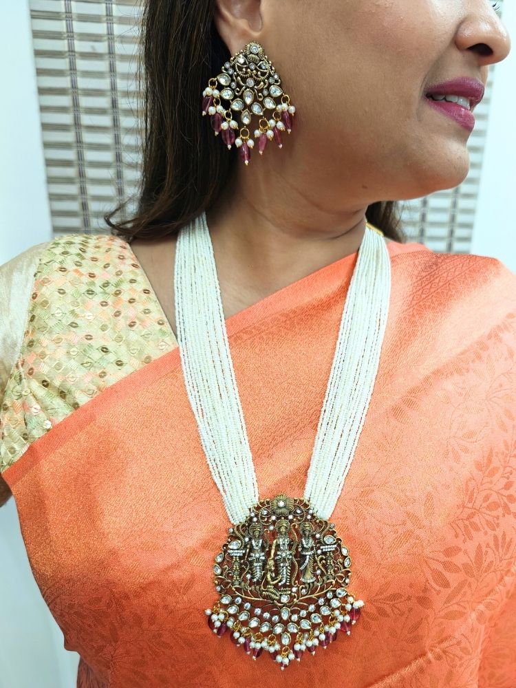 A woman smiles and looks to the side, wearing a Orange and gold saree with the Pink Rama Necklace Set—an ornate Kundan necklace with intricate designs and matching American Diamond earrings. Perfect necklace to add to your temple necklace collection. 