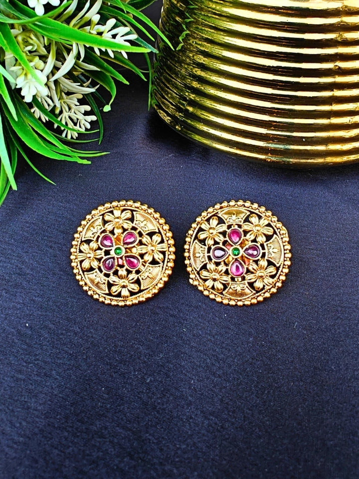 The Tarang Earrings are round, gold-toned studs featuring intricate floral designs and embedded Colored stones, showcased on a black fabric background near a gold ribbed container and green leaves.