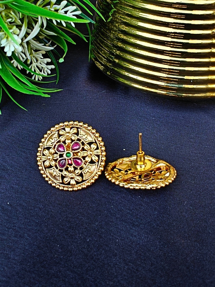 The Tarang Earrings are round, gold-toned studs featuring intricate floral designs and embedded Colored stones, showcased on a black fabric background near a gold ribbed container and green leaves.