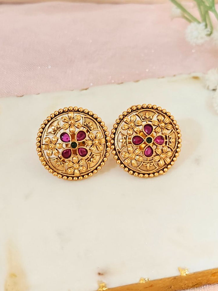 The Tarang Earrings are round, gold-toned studs featuring intricate floral designs and embedded Colored stones, showcased on a White Marble background with White Flower.