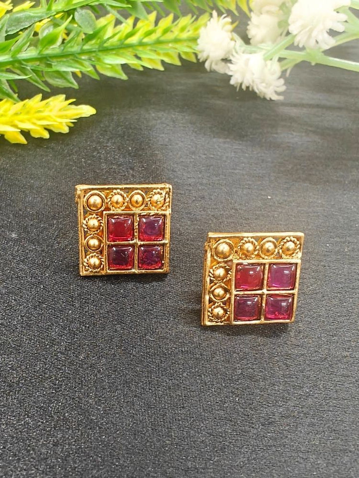 The Pink Vanita Earrings are square gold earrings with four red gemstones and gold bead detailing, shown on a black surface with yellow and white flowers—ideal for adding traditional flair to your Kurti ensemble.