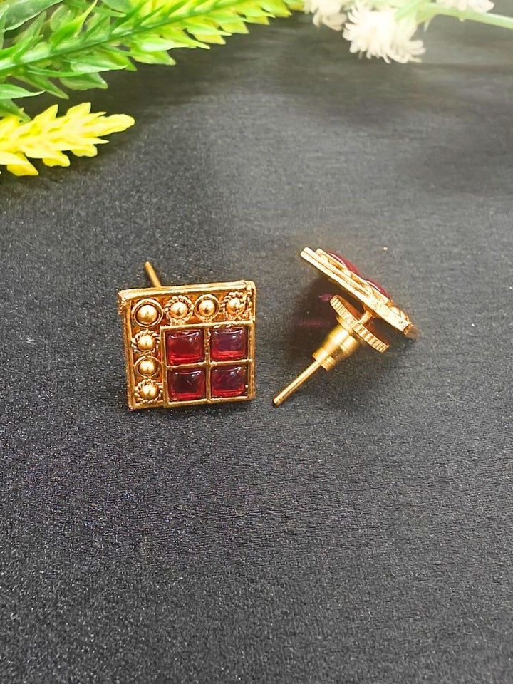 The Pink Vanita Earrings are square gold earrings with four red gemstones and gold bead detailing, shown on a black surface with yellow and white flowers—ideal for adding traditional flair to your Kurti ensemble.