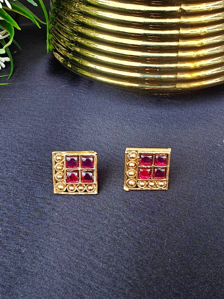 The Pink Vanita Earrings are square gold earrings with four red gemstones and gold bead detailing, shown on a black surface with yellow and white flowers—ideal for adding traditional flair to your Kurti ensemble.