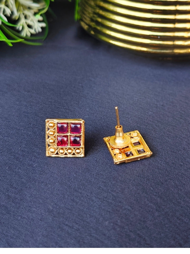 The Pink Vanita Earrings are square gold earrings with four red gemstones and gold bead detailing, shown on a black surface with yellow and white flowers—ideal for adding traditional flair to your Kurti ensemble.