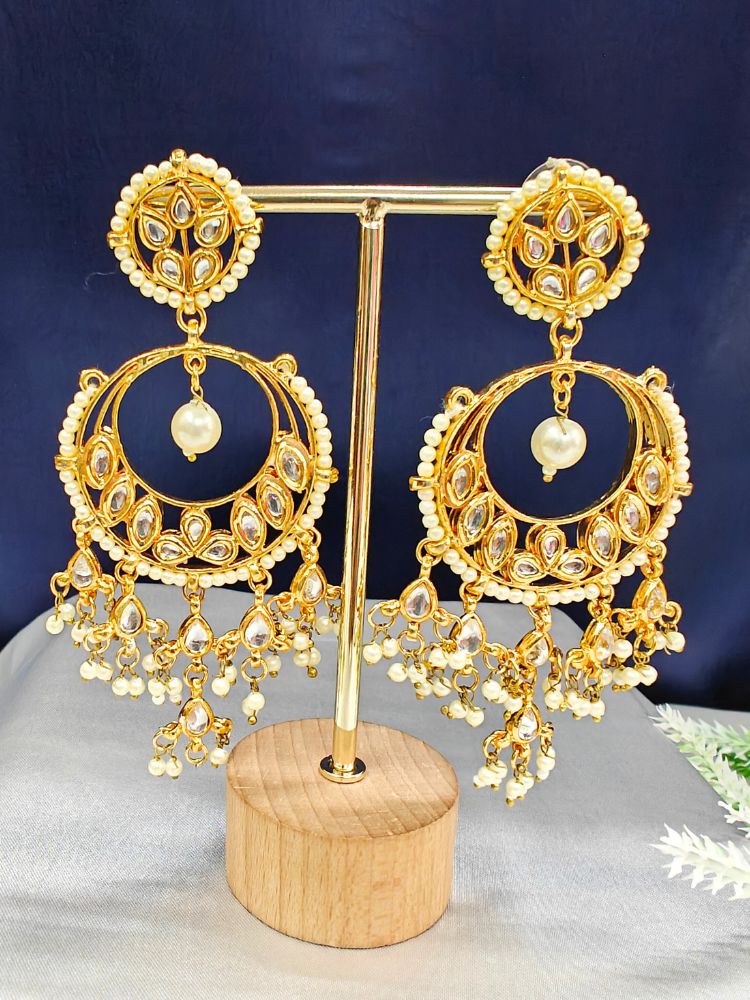 The Prabha Earrings are ornate gold chandelier kundan earrings with pearl accents and intricate kundan detailing, displayed on a gold stand with wooden base—perfect to complement your wedding ceremony dress.