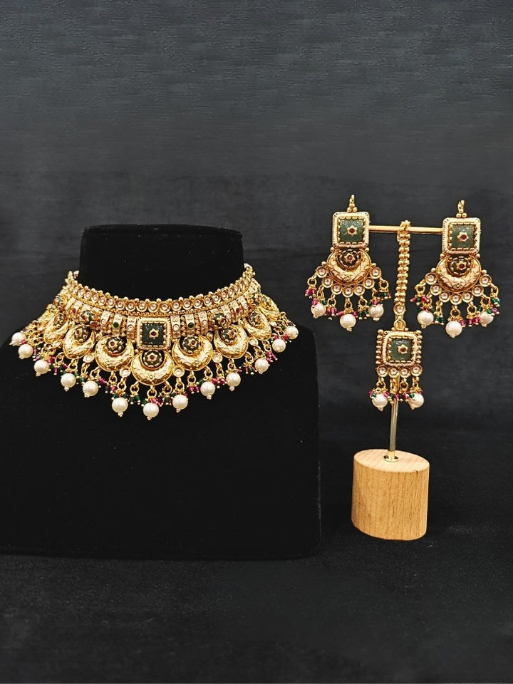 Close-up of an ornate traditional Rajwadi and Heritage gold Choker jewellery set and earrings and maangtikka with Pearls, Beads, Meenakari and Green Colored Stones. Close-up of a premium Rajwadi artificial necklace under 10000. Wedding guest styling a Rajwadi artificial necklace. Luxury wedding styling featuring an antique-inspired artificial Heritage necklace. Close-up of enamel work in Meenakari artificial wedding jewelry.