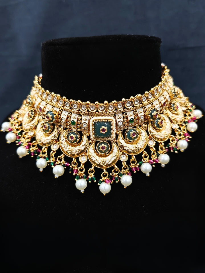 Close-up of an ornate traditional Rajwadi and Heritage gold Choker jewellery set and earrings and maangtikka with Pearls, Beads, Meenakari and Green Colored Stones. Close-up of a premium Rajwadi artificial necklace under 10000. Wedding guest styling a Rajwadi artificial necklace. Luxury wedding styling featuring an antique-inspired artificial Heritage necklace. Close-up of enamel work in Meenakari artificial wedding jewelry.