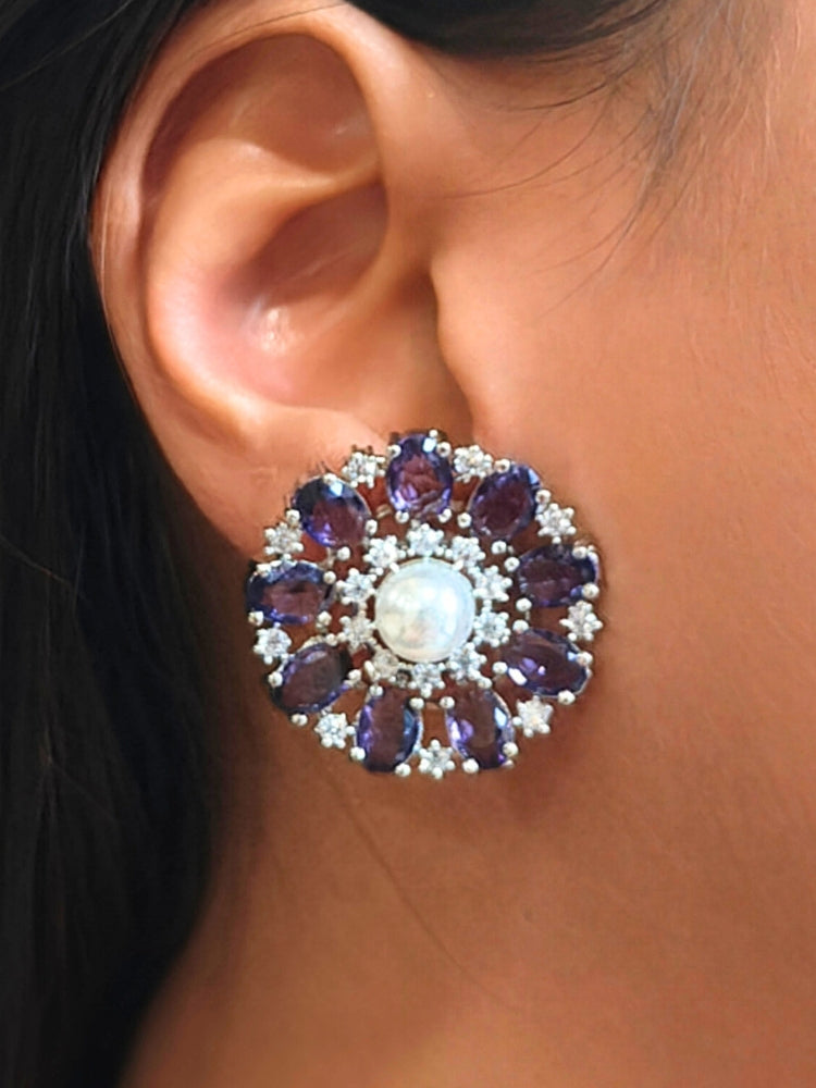 A close-up of a womans ear wearing the Purple Blossom stud Earrings—large, circular purple earrings with a pearl at the center, colorful accents, and a border of sparkling American Diamonds. The stud earrings give a flower earrings feel. 