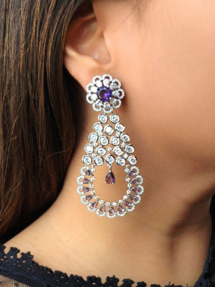 A close-up of a womans ear features the Purple Darcie Earrings, designed with American Diamonds in floral and circular patterns and finished with a dangling Purple teardrop gem—ideal for complementing a cocktail gown.
