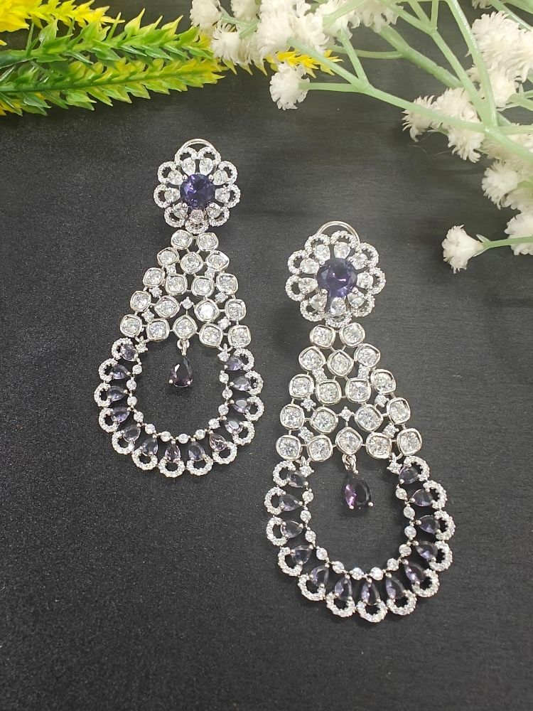 the Purple Darcie Earrings, designed with American Diamonds in floral and circular patterns and finished with a dangling Purple teardrop gem—ideal for complementing a cocktail gown. Its shown on a Black Surface with white flowers & Green, Yellow Leaves.