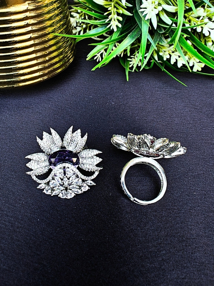 Two ornate, flower-shaped cocktail rings rest on a dark surface; one is the Purple Megan Ring, featuring Purple stones and sparkling American Diamond accents. The other ring is shown from behind, with green foliage and a gold object in the background.