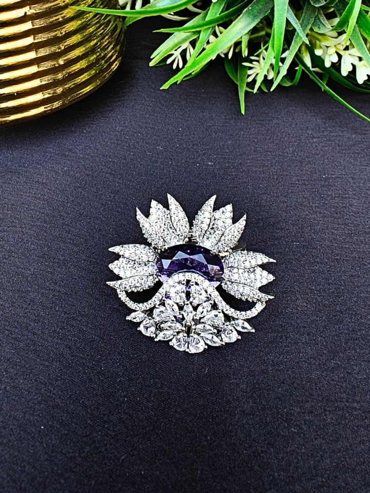 flower-shaped cocktail rings rest on a dark surface; this is the Purple Megan Ring, featuring Purple stones and sparkling American Diamond accents. The ring is shown with green foliage and a gold object in the background.