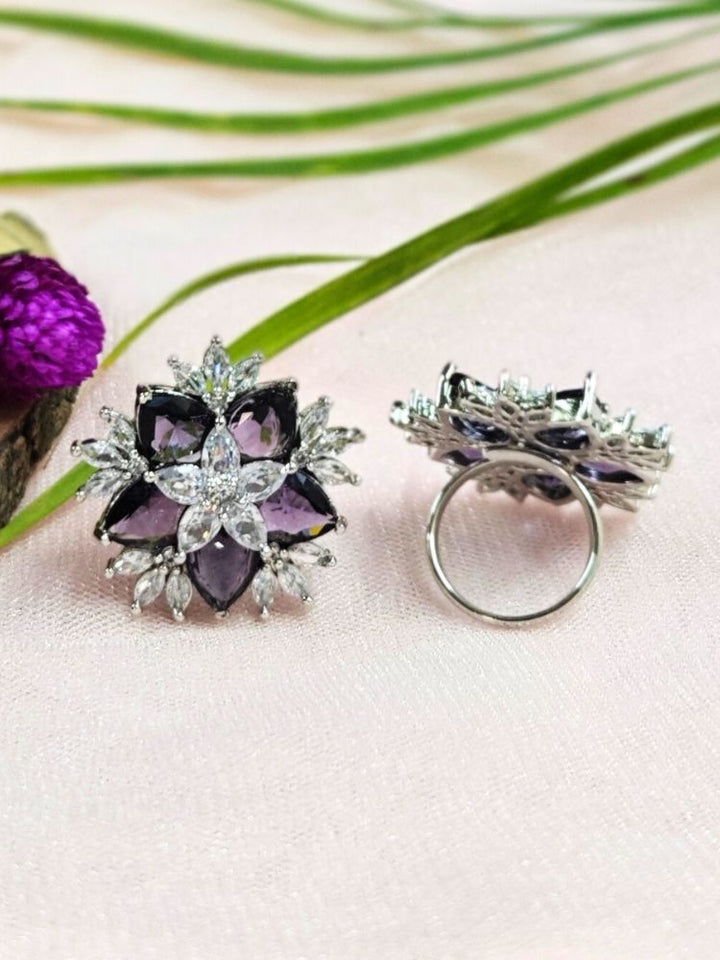 Two ornate, adjustable Purple Snowflake Rings with large purple gemstone and american diamonds in floral designs—ideal as cocktail rings—are displayed on a light pink surface with green leaves and a purple flower nearby.