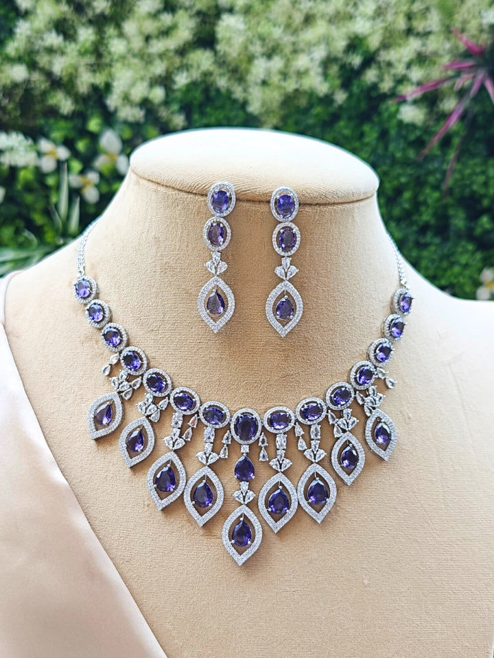 Purple Valerie Necklace Set, featuring an ornate silver necklace and matching earrings with pruple stones and sparkling American diamonds, shown on a cream dummy with greenery behind. 