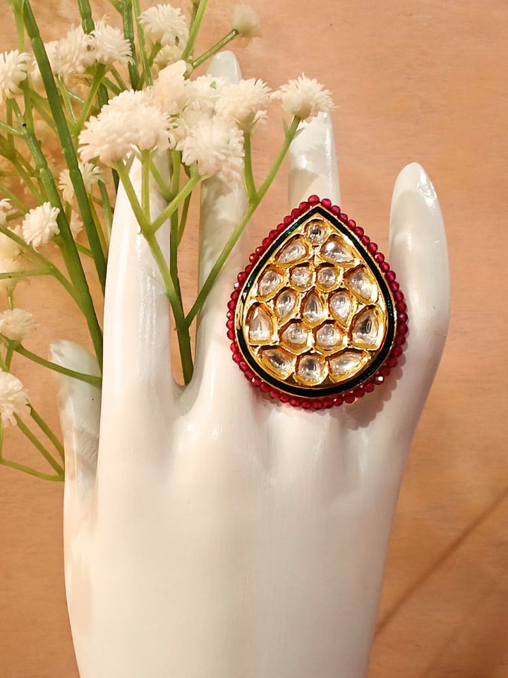 The Quin Ring, a large teardrop-shaped adjustable ring with gold and Kundan stones bordered by red crystal stones, is displayed on a white mannequin hand with white flowers in the background.