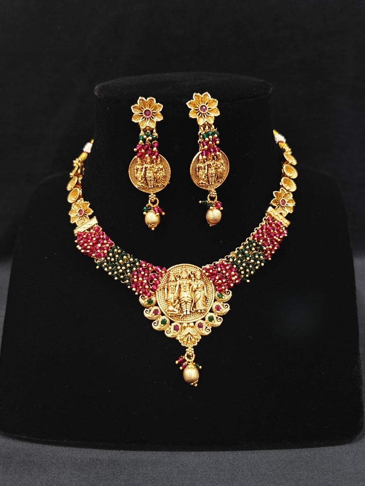 The Raghav Necklace Set featuring a traditional gold Collar Red & Green necklace and matching earrings with Goddess Ram in the middle, along with Red and green Colored Stone and Cyrstal Beads. Close-up of a temple imitation necklace styled with a silk saree. Festive Diwali look completed with a traditional temple necklace set.