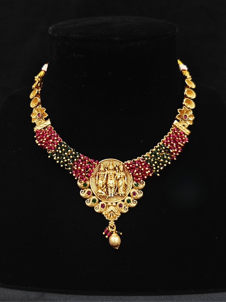 The Raghav Necklace Set featuring a traditional gold Collar Red & Green necklace and matching earrings with Goddess Ram in the middle, along with Red and green Colored Stone and Cyrstal Beads. Close-up of a temple imitation necklace styled with a silk saree. Festive Diwali look completed with a traditional temple necklace set.