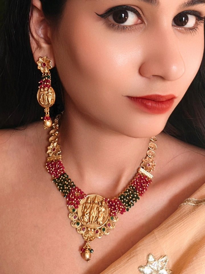 A woman wearing the Raghav Necklace Set featuring a traditional gold Collar Red & Green necklace and matching earrings with Goddess Ram in the middle, along with Red and green Colored Stone and Cyrstal Beads. Close-up of a temple imitation necklace styled with a silk saree. Festive Diwali look completed with a traditional temple necklace set.