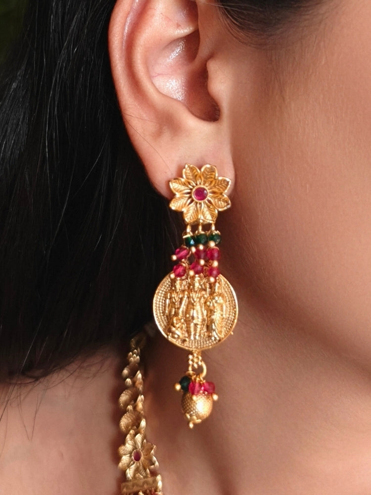 A woman wearing the Raghav Necklace Set featuring a traditional gold Collar Red & Green necklace and matching earrings with Goddess Ram in the middle, along with Red and green Colored Stone and Cyrstal Beads. Close-up of a temple imitation necklace styled with a silk saree. Festive Diwali look completed with a traditional temple necklace set.
