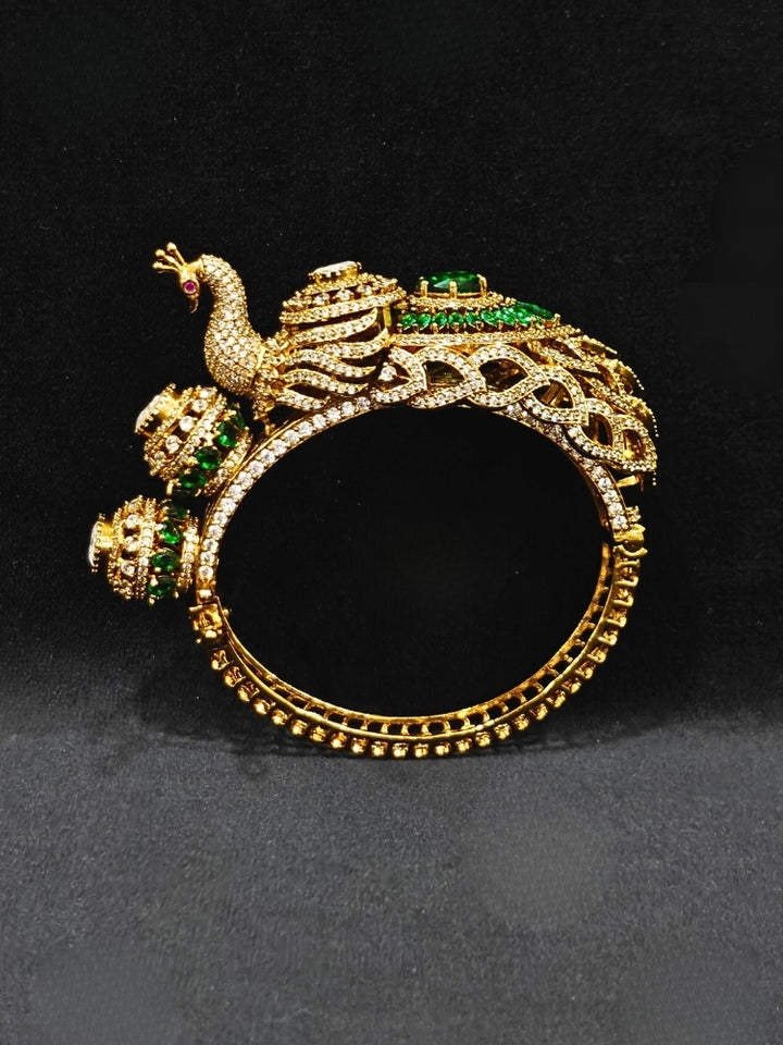 A close-up of the Rajmor Green Bracelet (Openable), featuring ornate gold work, a detailed peacock design with intricate patterns made with sparkling American diamonds and  green colored stones,  worn elegantly on the wrist—perfect as wedding jewelry.