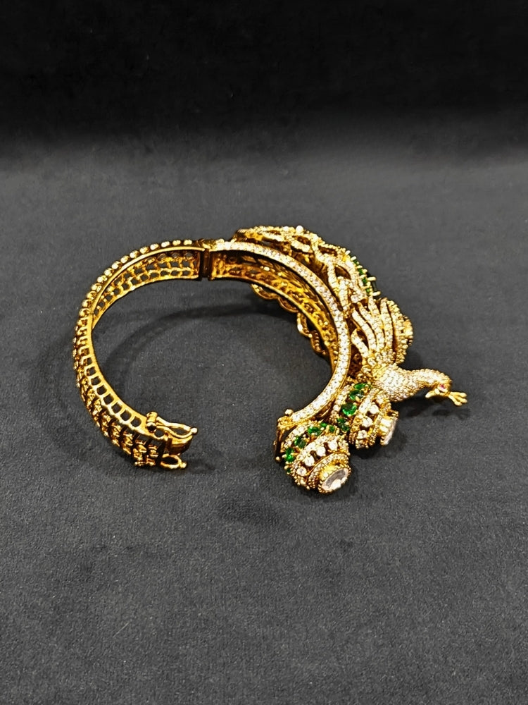 A close-up of the Rajmor Green Bracelet (Openable), featuring ornate gold work, a detailed peacock design with intricate patterns made with sparkling American diamonds and  green colored stones,  worn elegantly on the wrist—perfect as wedding jewelry.