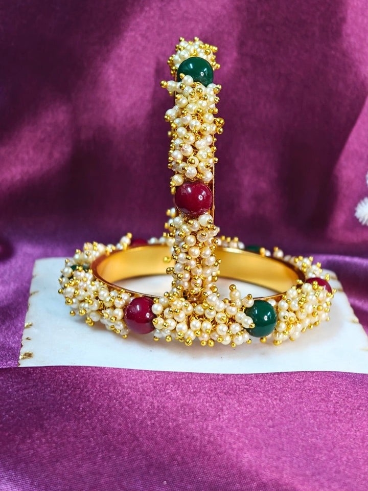 Close-up of a woman wearing an ornate traditional (Gold Polish) White, Red & Green Bangles set with Pearls and Colored Stones. Close-up of handcrafted artificial Pearl bangles with detailed design. Festive Navratri outfit paired with stacked imitation Pearl bangles.