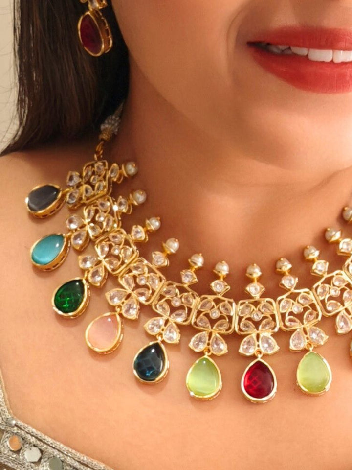 A woman radiates traditional elegance wearing the Rani Kundan Necklace Set with multi-color stones, complete with matching earrings. She smiles softly in an ornate, embroidered outfit accented by mirror embellishments.