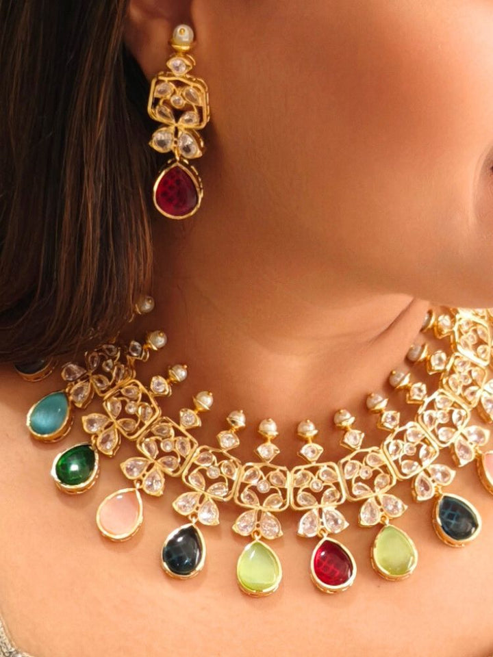 A woman radiates traditional elegance wearing the Rani Kundan Necklace Set with multi-color stones, complete with matching earrings. She smiles softly in an ornate, embroidered outfit accented by mirror embellishments.