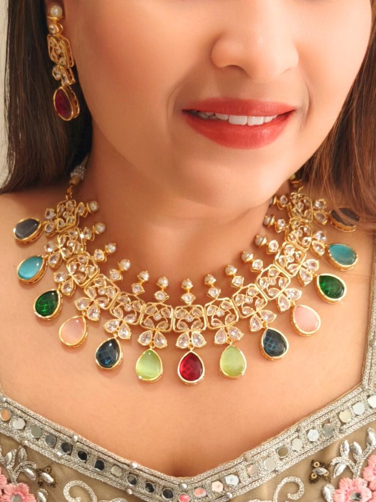 A woman radiates traditional elegance wearing the Rani Kundan Necklace Set with multi-color stones, complete with matching earrings. She smiles softly in an ornate, embroidered outfit accented by mirror embellishments.