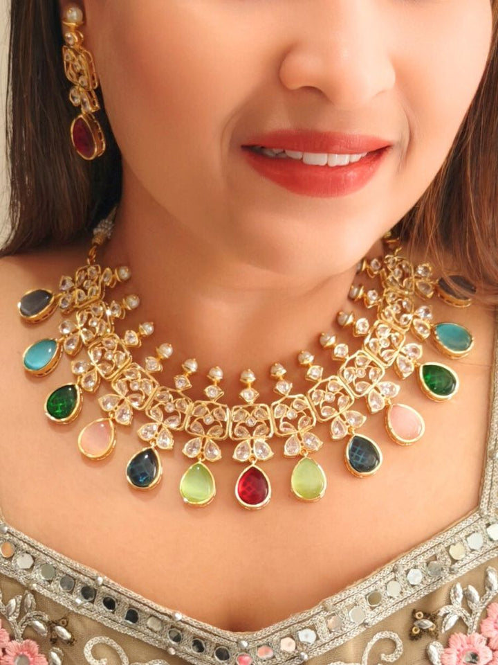 A woman radiates traditional elegance wearing the Rani Kundan Necklace Set with multi-color stones, complete with matching earrings. She smiles softly in an ornate, embroidered outfit accented by mirror embellishments.