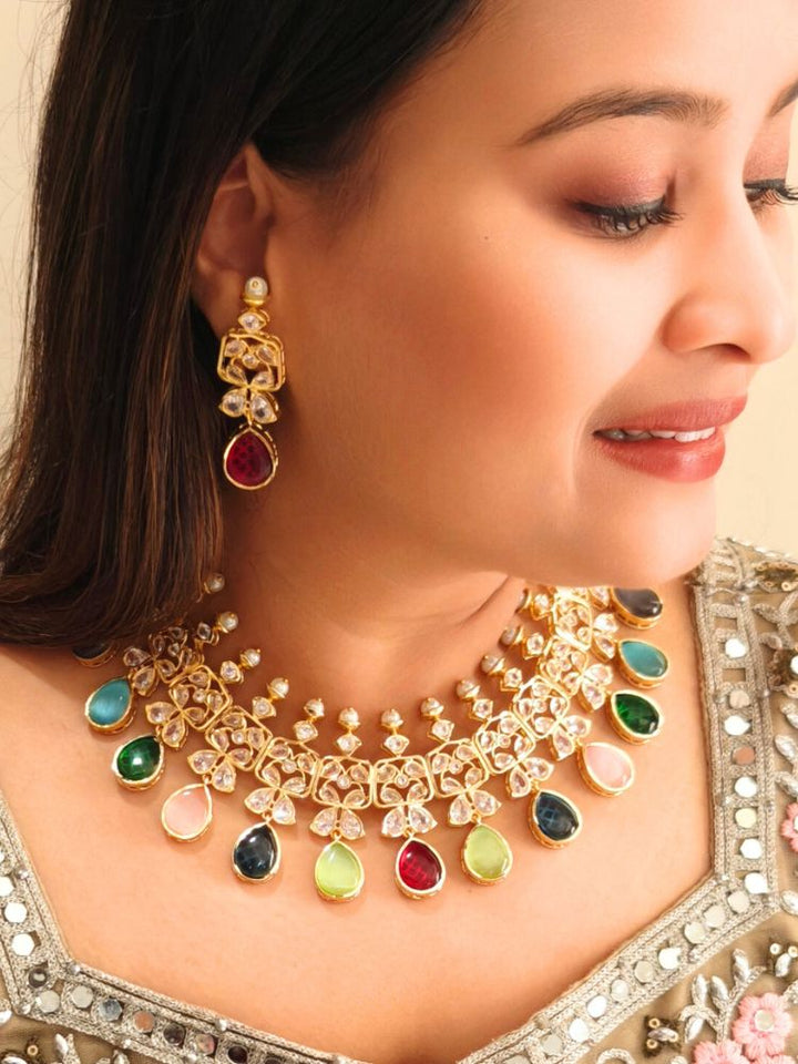 A woman radiates traditional elegance wearing the Rani Kundan Necklace Set with multi-color stones, complete with matching earrings. She smiles softly in an ornate, embroidered outfit accented by mirror embellishments.