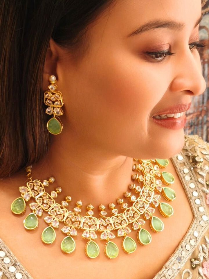A woman radiates traditional elegance wearing the Rani Kundan Necklace Set with Parrot Green stones, complete with matching earrings. She smiles softly in an ornate, embroidered outfit accented by mirror embellishments.