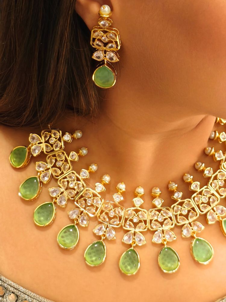 A woman radiates traditional elegance wearing the Rani Kundan Necklace Set with Parrot Green stones, complete with matching earrings. She smiles softly in an ornate, embroidered outfit accented by mirror embellishments.