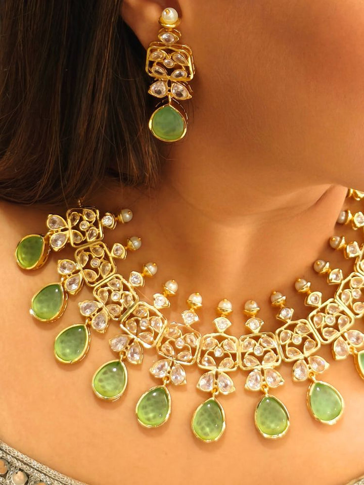 A woman radiates traditional elegance wearing the Rani Kundan Necklace Set with Parrot Green stones, complete with matching earrings. She smiles softly in an ornate, embroidered outfit accented by mirror embellishments.