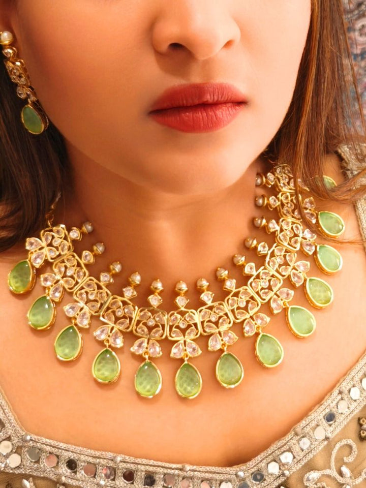 A woman radiates traditional elegance wearing the Rani Kundan Necklace Set with Parrot Green stones, complete with matching earrings. She smiles softly in an ornate, embroidered outfit accented by mirror embellishments.