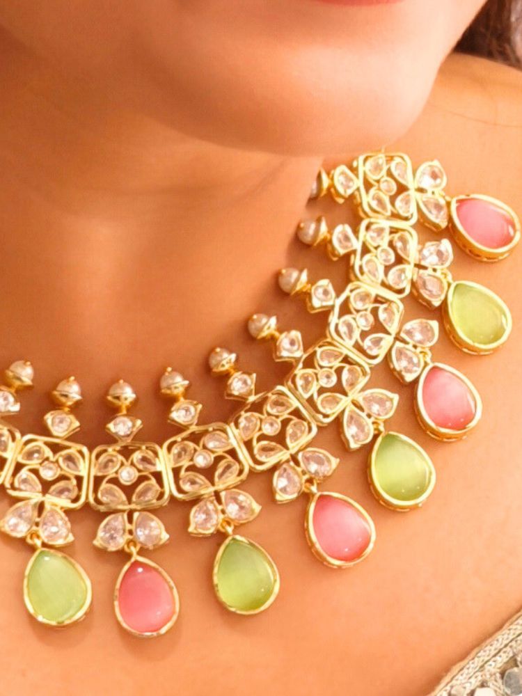 A woman radiates traditional elegance wearing the Rani Kundan Necklace Set with Pink & Green stones, complete with matching earrings. She smiles softly in an ornate, embroidered outfit accented by mirror embellishments.
