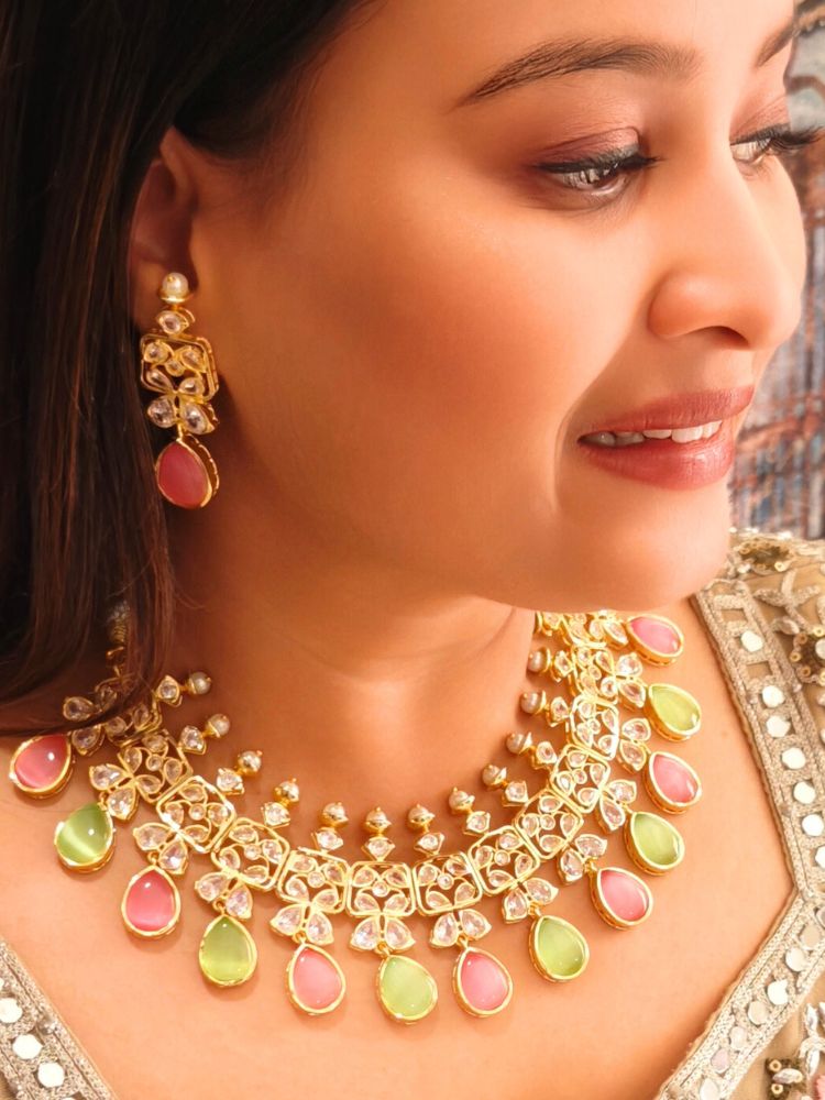 A woman radiates traditional elegance wearing the Rani Kundan Necklace Set with Pink & Green stones, complete with matching earrings. She smiles softly in an ornate, embroidered outfit accented by mirror embellishments.