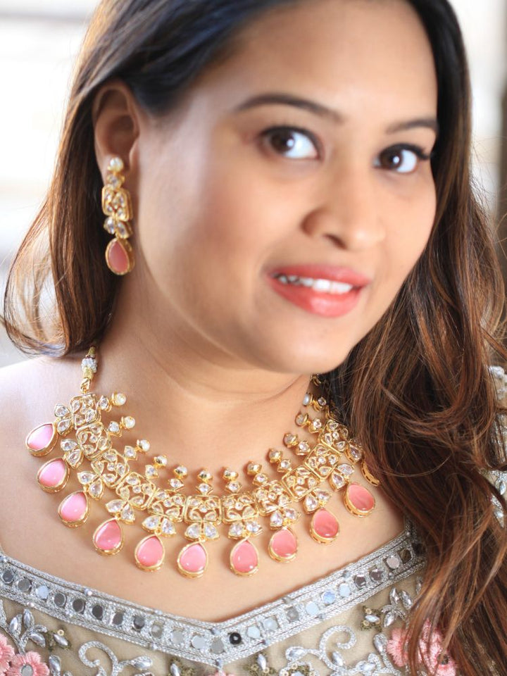 A woman radiates traditional elegance wearing the Rani Kundan Necklace Set with Pink stones, complete with matching earrings. She smiles softly in an ornate, embroidered outfit accented by mirror embellishments.