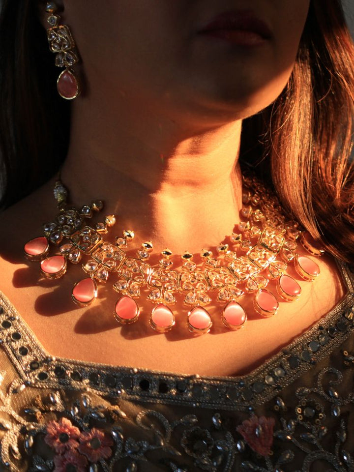 A woman radiates traditional elegance wearing the Rani Kundan Necklace Set with Pink stones, complete with matching earrings. She smiles softly in an ornate, embroidered outfit accented by mirror embellishments.