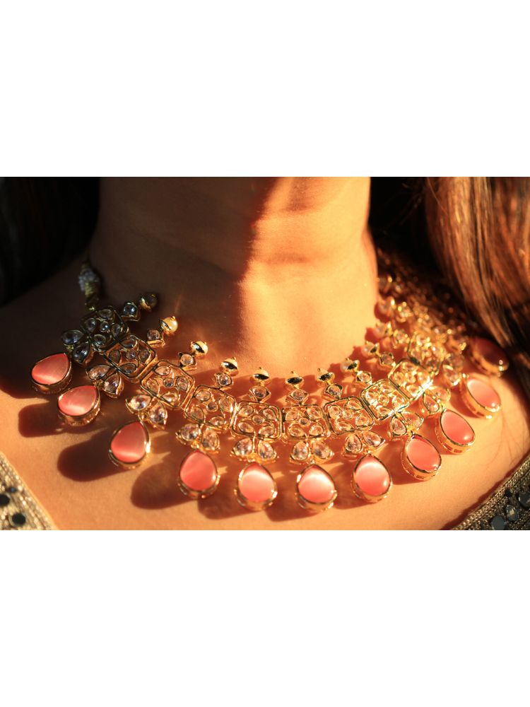 A woman radiates traditional elegance wearing the Rani Kundan Necklace Set with Pink stones, complete with matching earrings. She smiles softly in an ornate, embroidered outfit accented by mirror embellishments.