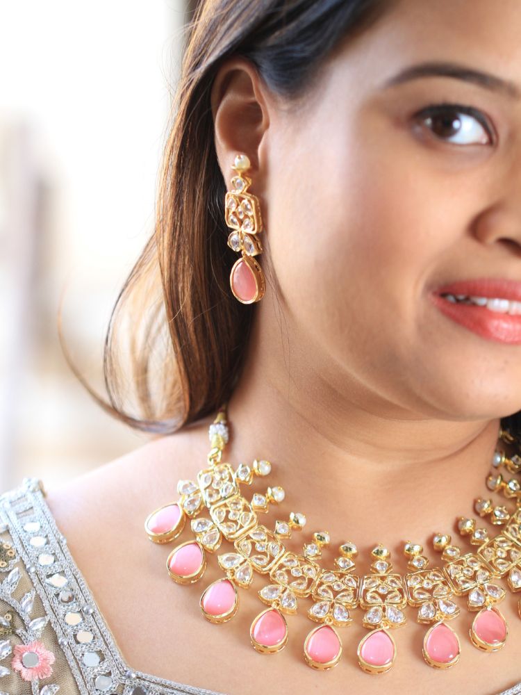 A woman radiates traditional elegance wearing the Rani Kundan Necklace Set with Pink stones, complete with matching earrings. She smiles softly in an ornate, embroidered outfit accented by mirror embellishments.