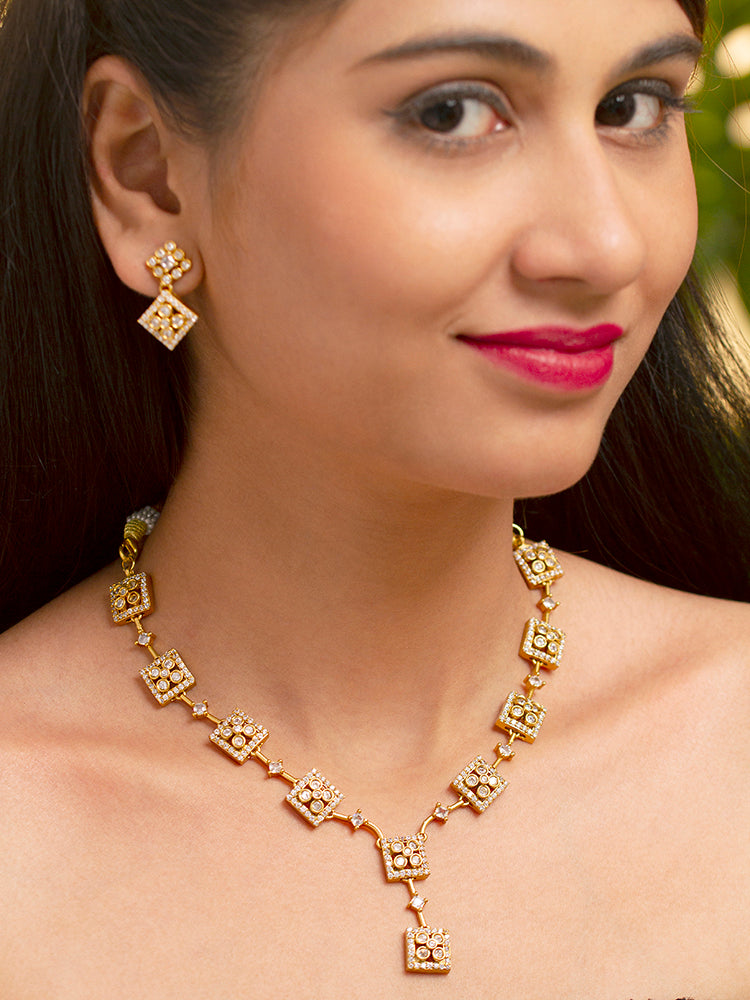 A woman with long dark hair wears the Ravira Necklace Set—gold earrings and a matching gold Y- shape necklace with square designs and intricate patterns. She smiles gently and is dressed in traditional attire.