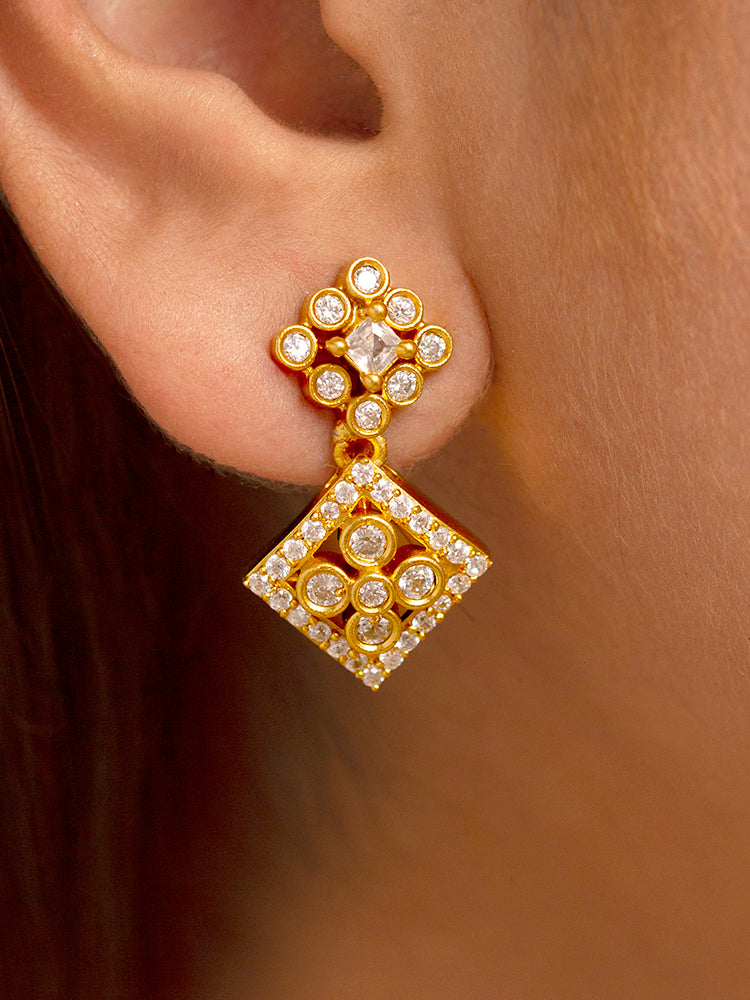 A woman with long dark hair wears the Ravira Necklace Set—gold earrings and a matching gold Y- shape necklace with square designs and intricate patterns. She smiles gently and is dressed in traditional attire.