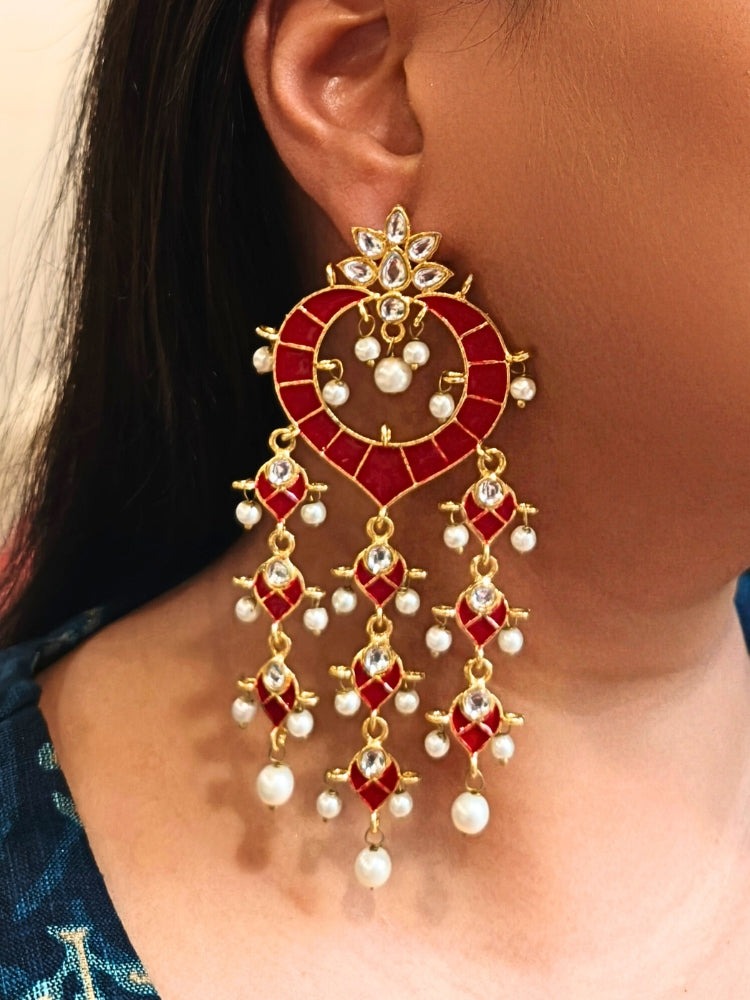 A close-up of a person wearing Red Ankita Earrings—large, ornate Red and gold earrings with dangling pearls and gemstones in intricate patterns—standing out against dark hair and a blue patterned top. It has meenakari design all over the earrings, and is perfect for wedding functions. 