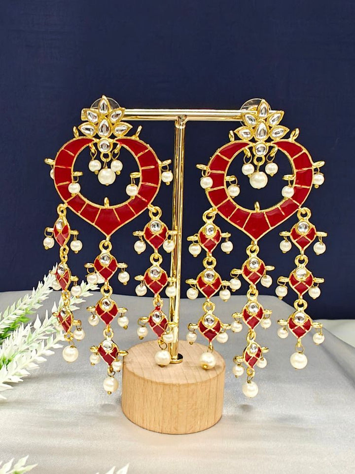 The Red Ankita Earrings are ornate gold Kundan earrings with Red enamel, intricate Meenakari work, white stones, and dangling pearl beads, shown on a gold stand with a wooden base against a dark background.