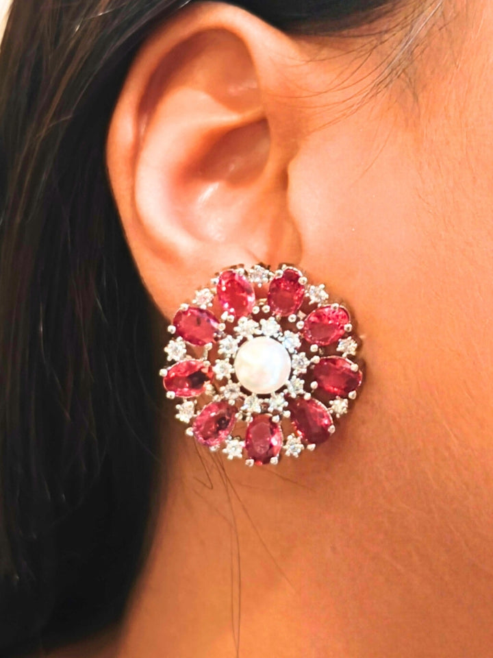 A close-up of a womans ear wearing the Red Blossom stud Earrings—large, circular Red earrings with a pearl at the center, colorful accents, and a border of sparkling American Diamonds. The stud earrings give a flower earrings feel. 