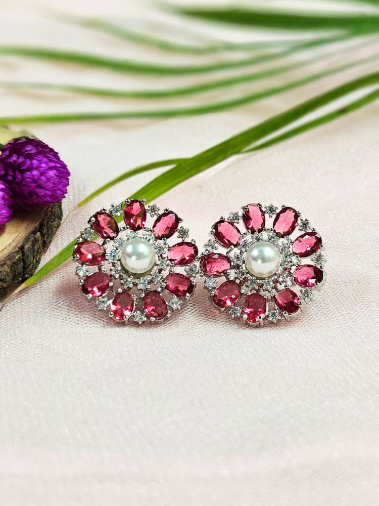 The Red Blossom Earrings showcase a central white pearl encircled by alternating Red stones and American diamonds, elegantly displayed on soft pink fabric with green leaves and a purple flower.