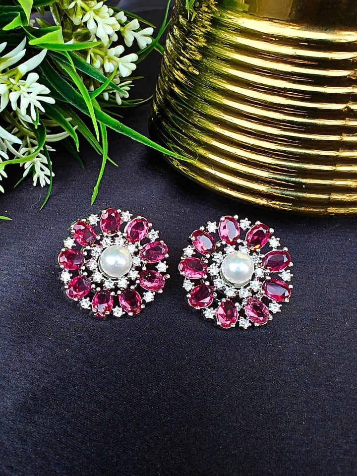 The Red Blossom Earrings showcase a central white pearl encircled by alternating Red stones and American diamonds, elegantly displayed on Black fabric with green leaves and a Golden accent.
