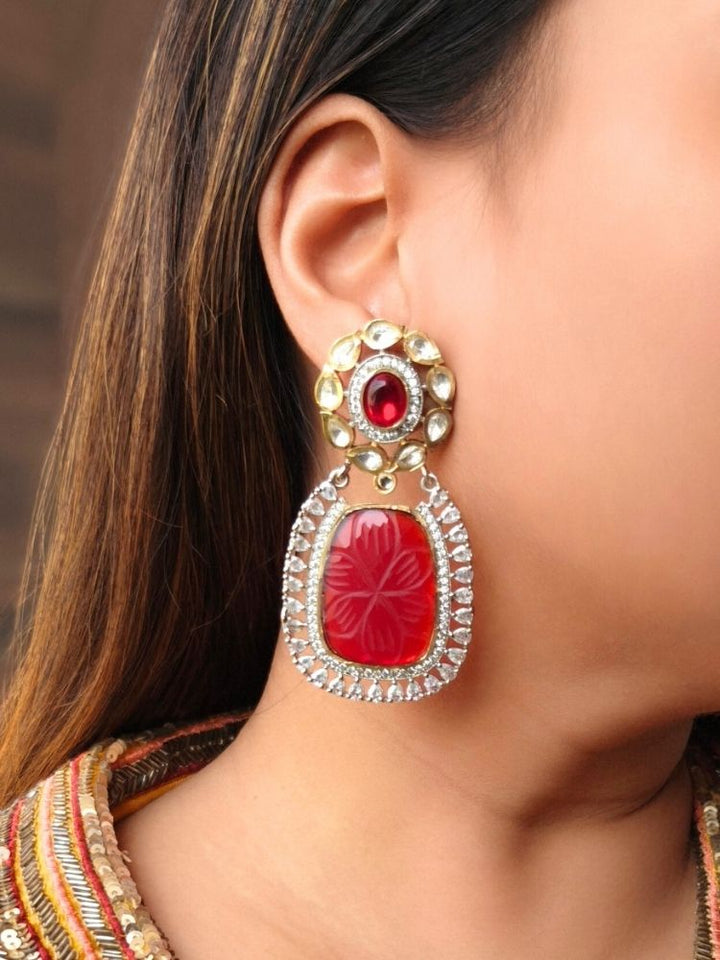 A close-up of a womans ear wearing Ruby Red Fiona Earrings featuring an Ruby Red carved stone, ornate American Diamonds and Kundan. Her dark hair is partially visible, and she wears a colorful embroidered garment.