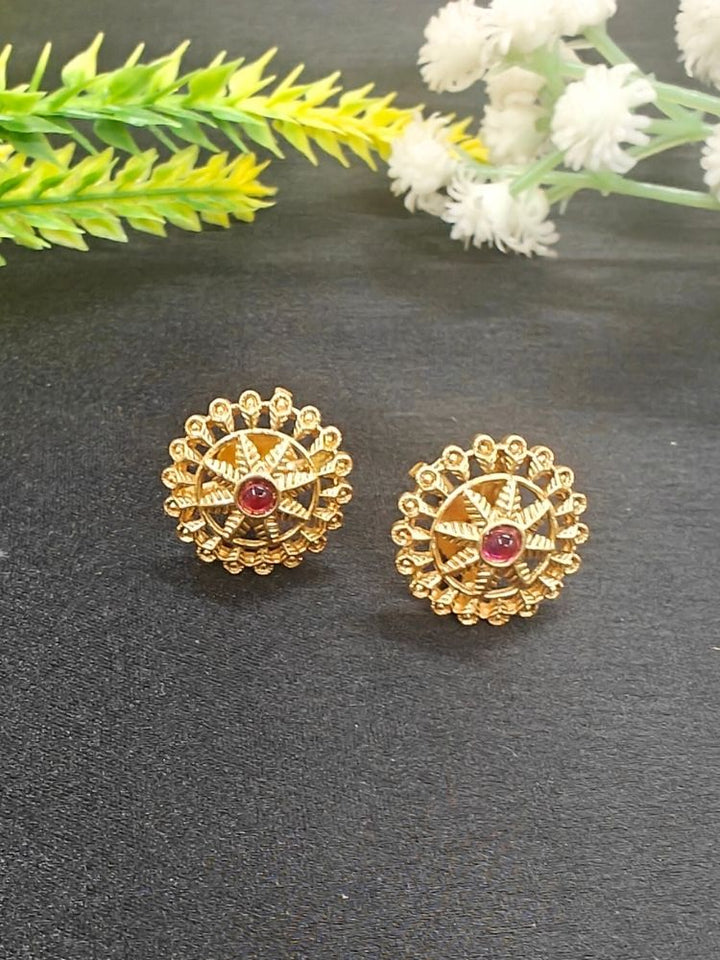 The Red Gehna Earrings are traditional round gold stud earrings with intricate designs and a central ruby-colored stone, displayed on a dark surface with white and yellow artificial flowers in the background.